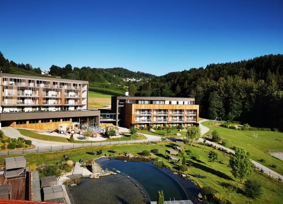 Familienhotel Schreinerhof im Bayerischen Wald: Hotelgebäude in Holzarchitektur mit Naturbadeteich, Spielwiese und Wald im Hintergrund