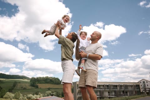 Eltern heben ihre beiden Kleinkinder lachend in den Himmel, im Hintergrund das Hotel