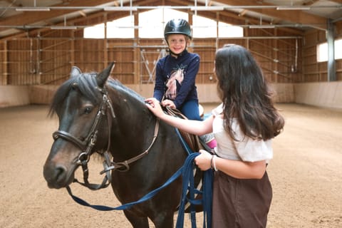 Kind mit Reithelm sitzt auf einem Pony in der Reithalle, eine Frau hält das Pony