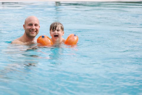 Papa und Kleinkind mit orangen Schwimmflügeln lachen gemeinsam im Pool