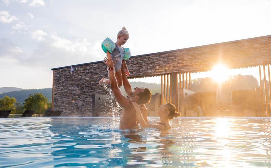 Familie im Außenpool des Familienhotels Schreinerhof, Vater hebt sein Kind aus dem Wasser, im Hintergrund die Abendsonne